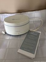 Closed white LaSonic Connoisseurs sonic jewellery cleaner unit on a cloth surface with the instruction manual beside it.