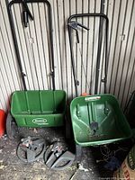 Photo of Scotts seed spreader and another green plastic spreader with wheels and handles along with two circular sprinkler heads on the floor.