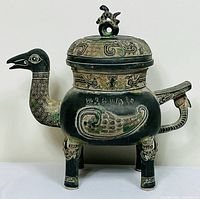 Front and side view of bronze bird-shaped pot with clear view of body, legs, handle, spout, and lid with finial. Shows detailed engraving and patina.