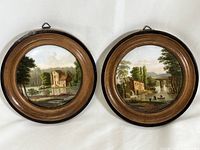 Two framed porcelain plates with hand-painted rural landscapes showing buildings, trees, and water. Mounted in round wooden frames with brass hangers.