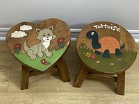 Two wooden stools side by side on a wooden floor; left stool is heart-shaped with a cat carving and painting; right stool is round with a tortoise carving and painting.