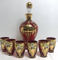 Front view of decanter and six matching small glasses, showing the red glass with gold plating and enamel flowers.