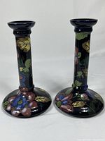 Pair of dark glazed pottery candlesticks decorated with Jacobean fruit, flower, and leaf motifs viewed from the front.