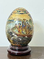 Full view of the hand painted Satsuma porcelain egg on wooden stand showing detailed river scene with figures.