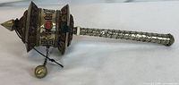 Side angle of Tibetan prayer wheel showing the drum with engraved Tibetan script, coral stones, pointed top finial, and chain with metal striker ball. Textured handle with scale-like pattern.