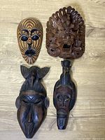 Four wooden masks displayed on a wooden floor: one wicker patterned mask, one carved mythical creature mask, two dark wood stylized faces with bird carvings.