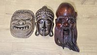 Three hand carved wooden masks arranged horizontally on floor showing distinct facial expressions and detailing.