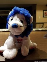 Close-up photo of the white plush lion with blue mane and embroidered number 99 on the chest, front view showing face and whiskers