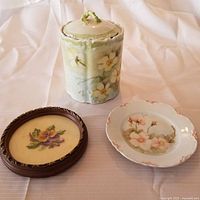 Photo showing the floral ceramic cookie jar with lid, small round framed petit point, and hand-painted floral plate on white fabric