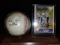 Photo of a white baseball in a clear display case featuring Nolan Ryan's stamped autograph and number 34 next to an autographed baseball card of Nolan Ryan in Texas Rangers uniform.