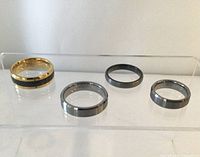 Four tungsten carbide rings displayed on clear stand, showing variety in color and design with one gold metallic and black ring plus three silver-gray metallic rings.