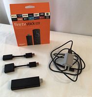 Amazon Fire TV Stick Lite box with power adapter, two HDMI extenders and streaming device laid out on white surface.