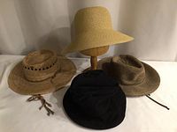 Four hats displayed together including a black corduroy vintage hat, natural straw hat, tan wide-brim hat on stand, and brown hat with chin strap.