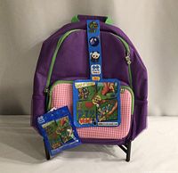 Front view of purple kids knapsack with pink pixel customizable front panel and included blue packet of pixel pieces.