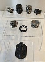 Eight stainless steel rings displayed on clear acrylic ring stand showing skulls, patterns, and plain band, plus black cobra dog tag pendant below.