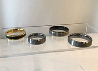 Four tungsten carbide rings displayed on clear acrylic stand showing different ring styles and finishes.