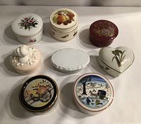 Eight porcelain and ceramic trinket boxes arranged in two rows, various shapes and decorative designs visible.