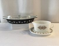 Front view of the two Pyrex items: matte black divided snowflake dish with clear lid and white gravy bowl with matching saucer with butterfly design.