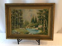 Full view of framed landscape painting with stand, showing wooded river scene with trees, rocks, and mountains in background.