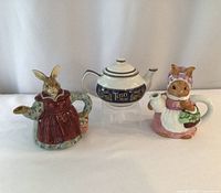 Three ceramic teapots side by side: rabbit in red dress with basket, classic Tetley Teas ceramic teapot blue and gold, rabbit in bonnet and pink dress with basket