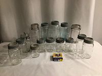 Front view of 16 vintage glass canning jars varying in size and lid type, presence of clear and blue tinted glass, arranged in two rows with a box of Velvety matches for scale.