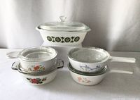 Full view of seven vintage ovenware pieces with various floral patterns and glass lids, arranged in two rows