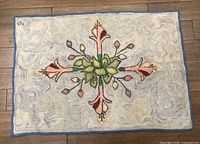Full top-down view of the hooked rug showing the entire floral pattern and blue border on hardwood floor.