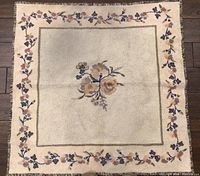 Full view of vintage handmade square rug with central floral motif and floral border on cream background.