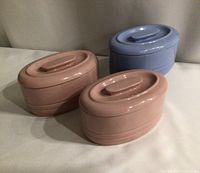 Three vintage oval refrigerator dishes with lids, two pink and one blue, displayed on a flat surface