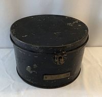 Front view of the round black metal hat tin with brass plaque and latch showing wear and paint loss.
