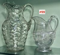 Two Victorian glass pitchers on display shelf with visible hand-applied handles and detailed body textures.
