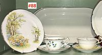 Two Shelley plates, one with a floral landscape, and two Royal Doulton platters in background, also two cups and saucers in front arranged on green surface.