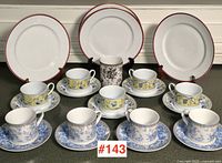 Full lot showing 4 white plates with red rim, multiple cups and saucers in blue and yellow floral patterns, and one black and white floral cup.