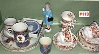 Assorted mugs with Royalty profile motifs, floral porcelain teacups stacked, and figurine of woman in blue