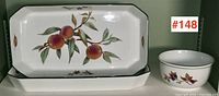 Two rectangular serving trays and one round bowl decorated with peaches and leaves arranged and photographed on a shelf against a green wall.
