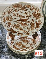 Three stacked plates showing painted bird, butterfly and insect motifs in brown and gilt on white porcelain.