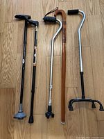 Photo showing five assorted canes standing on a wooden floor, including different styles like folding, wooden, and quad base canes.