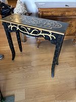 Full view of small black console table with gold decorative floral pattern and curved legs on wooden floor.