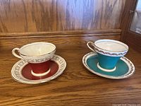 Two Aynsley teacups with matching saucers, one in red with gold design, the other in turquoise with gold detail.