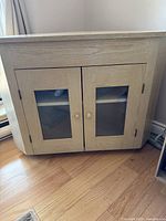 Front view of the wooden corner TV stand showing two glass paneled cupboard doors with wooden knobs.