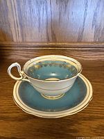 Teacup placed on saucer showing exterior design with turquoise blue band and gold floral detailing on white fine bone china