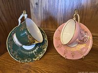 Green and pink Aynsley bone china teacups with matching saucers, gold floral detail and rims, displayed on wood shelf.