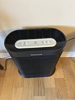 Black Honeywell air purifier standing on a wooden floor near a wall with power cord plugged in.
