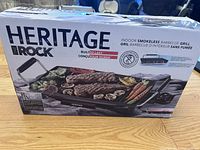 Box of Heritage The Rock Indoor Smokeless Barbecue Grill with branding and product image showing grill with food on surface and handle.