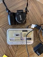 Front view of Kodak EasyShare DX7590 digital camera with lens cap on, camera strap visible, and Kodak docking station with power cord placed on a table.