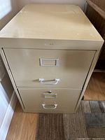 Front view of a beige metal filing cabinet with two drawers, each with a metal handle and label holder. Cabinet shows wear and minor damage.