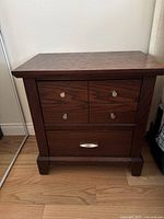 Front view of night table showing two small drawers on top with round metal knobs and one large drawer below with a metal handle. Dark wood grain visible.