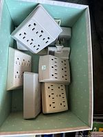 Box containing multiple white outlet splitters and power bars in rectangular and cube shapes with varying outlet counts.