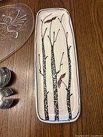 Rectangular ceramic serving platter with tree and cardinal bird design, clear glass oval serving tray with floral etching, and one stainless steel salad spoon with clear plastic handle, displayed on wooden surface.