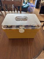 Closed yellow plastic sewing box with clear textured lid and white latch, sitting on wooden table